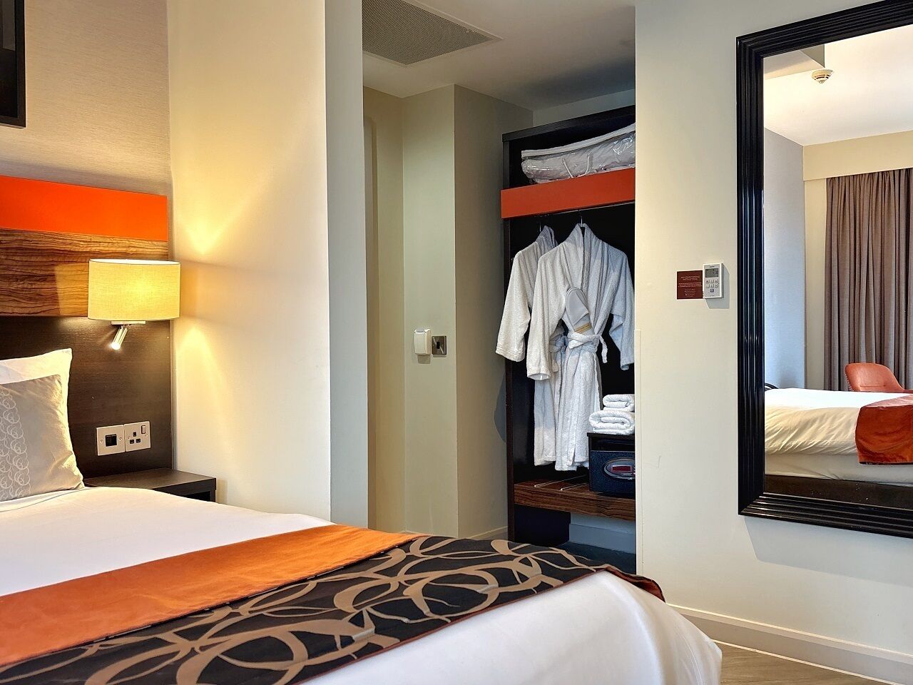 A hotel room with a bed, a nightstand, a lamp, a robe rack, and a mirror reflecting a bed and a chair.