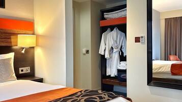 A hotel room with a bed, a nightstand, a lamp, a robe rack, and a mirror reflecting a bed and a chair.