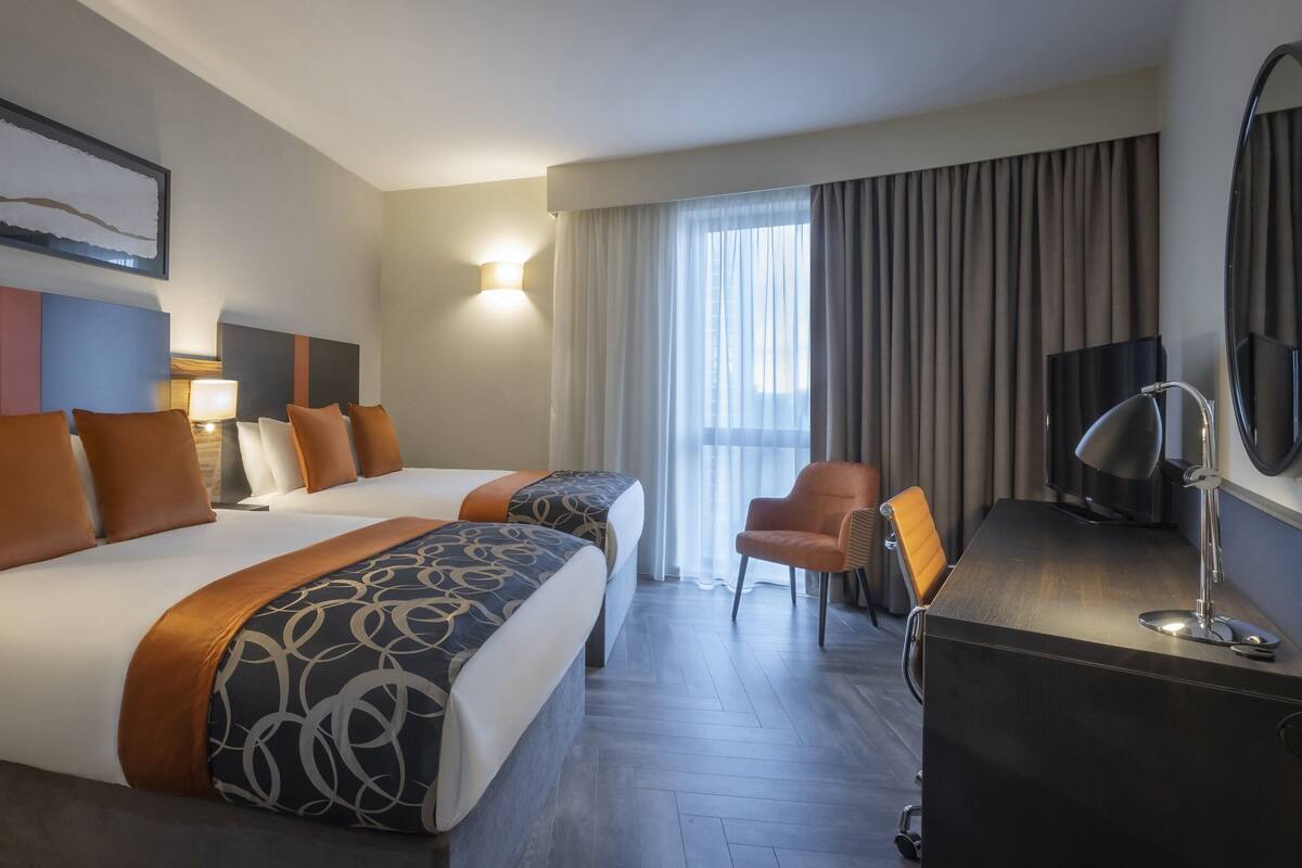 A hotel room with two beds, a desk with a chair, a TV, and a large window with curtains.