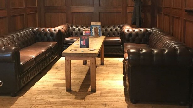 A room with wooden paneled walls, a wooden floor, a central table, and leather sofas arranged in a U-shape. - The Anchor Inn (March)