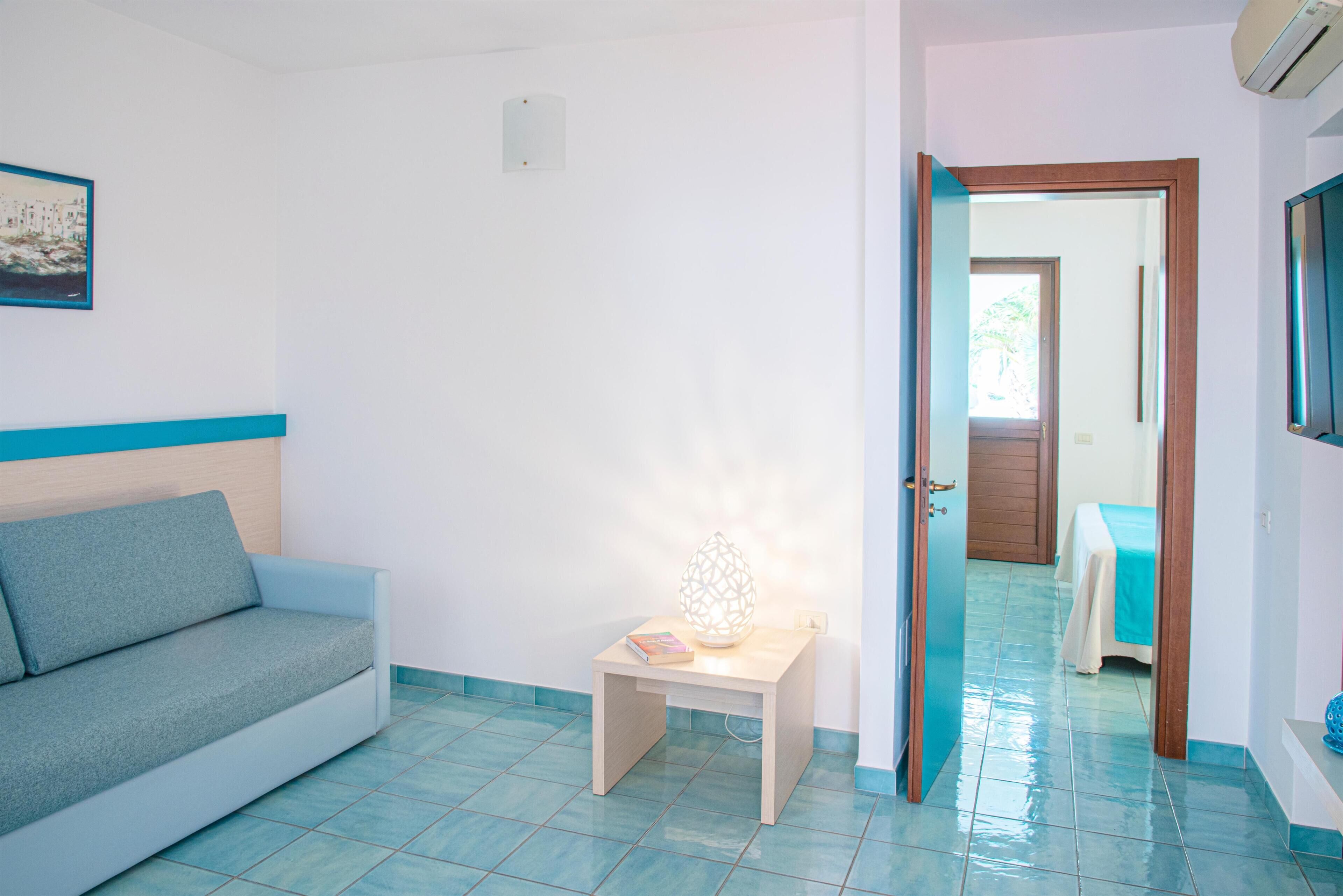 A room with a light blue tiled floor, a grey sofa, a small wooden table with a book and a decorative light, and a door leading to another room.