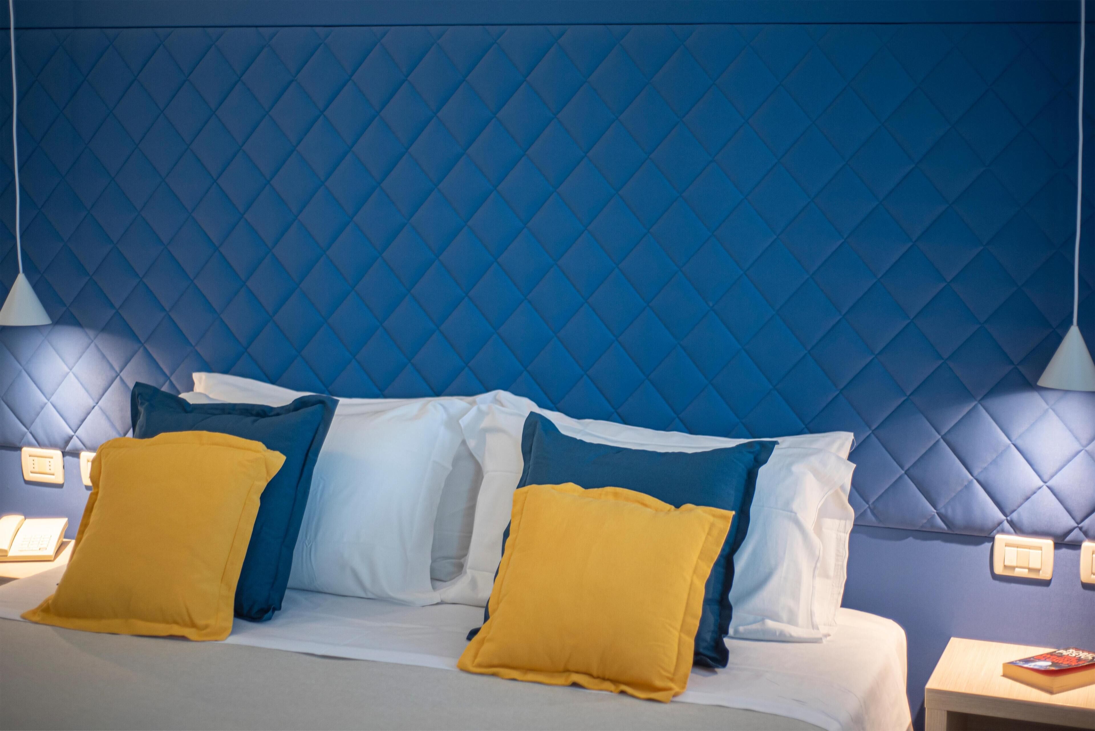 Una habitación de hotel con cabecera tapizada azul, almohadas blancas y amarillas, una mesita de noche con un libro y una lámpara.