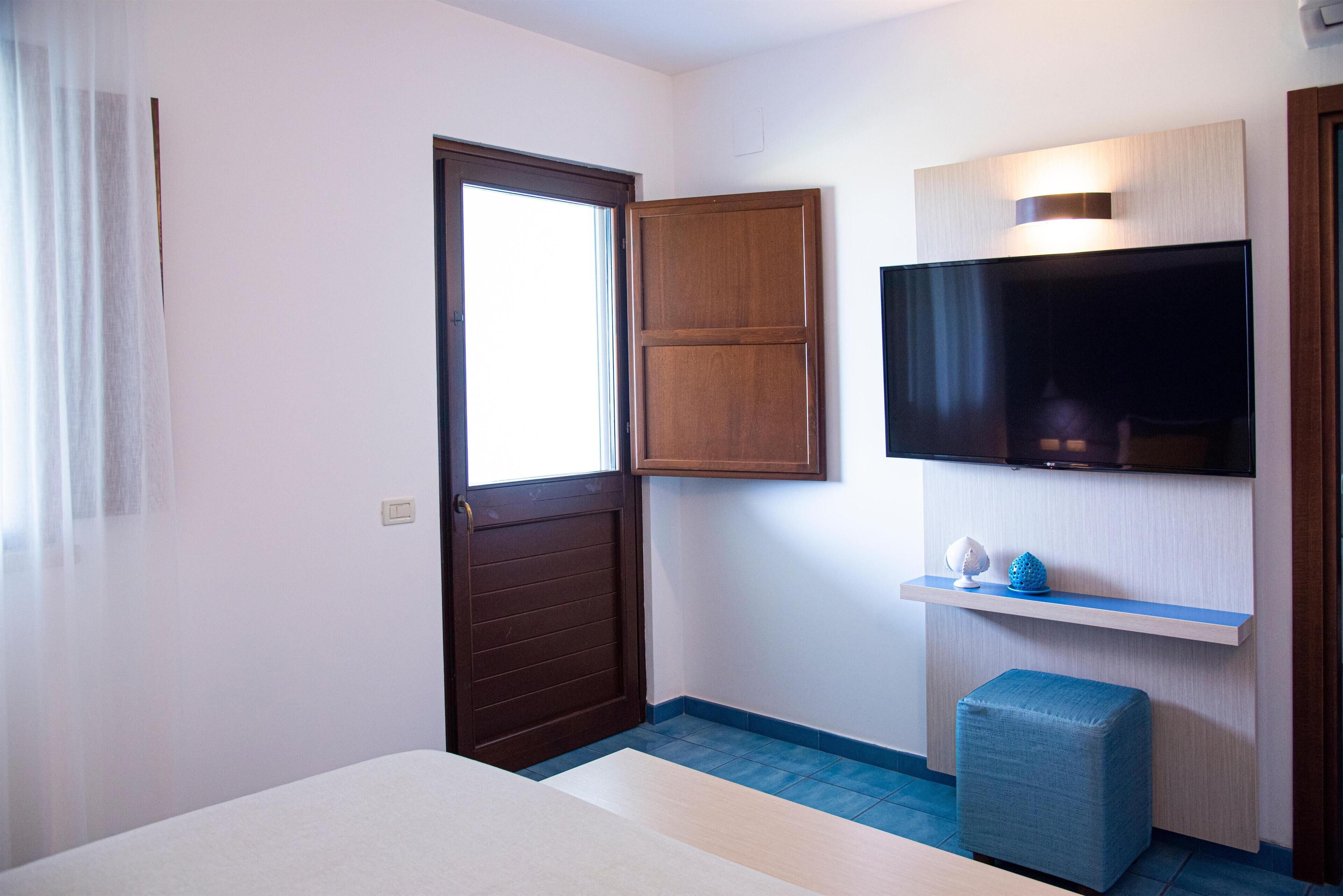 Camera d'albergo con un letto, una televisione fissata al muro, una porta in legno e un poggiapiedi blu.
