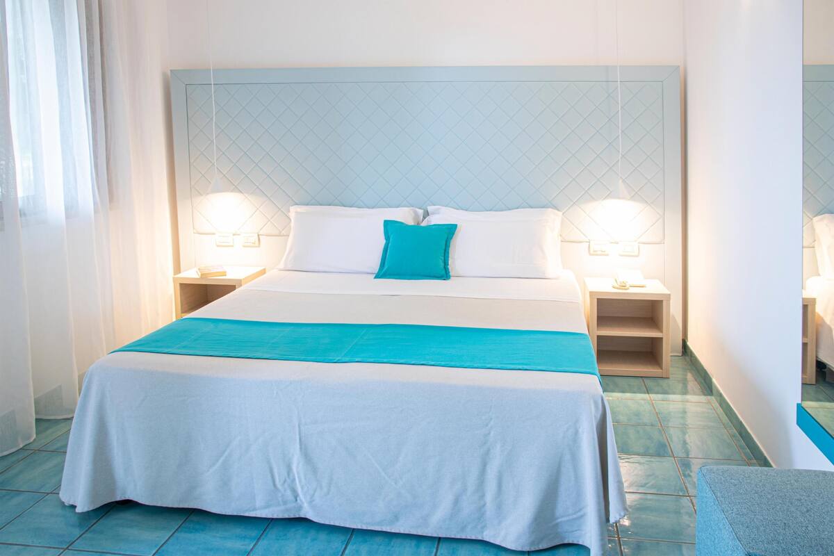 Quarto de hotel com uma cama grande, duas mesas de cabeceira e cabeceira com padrão acolchoado.