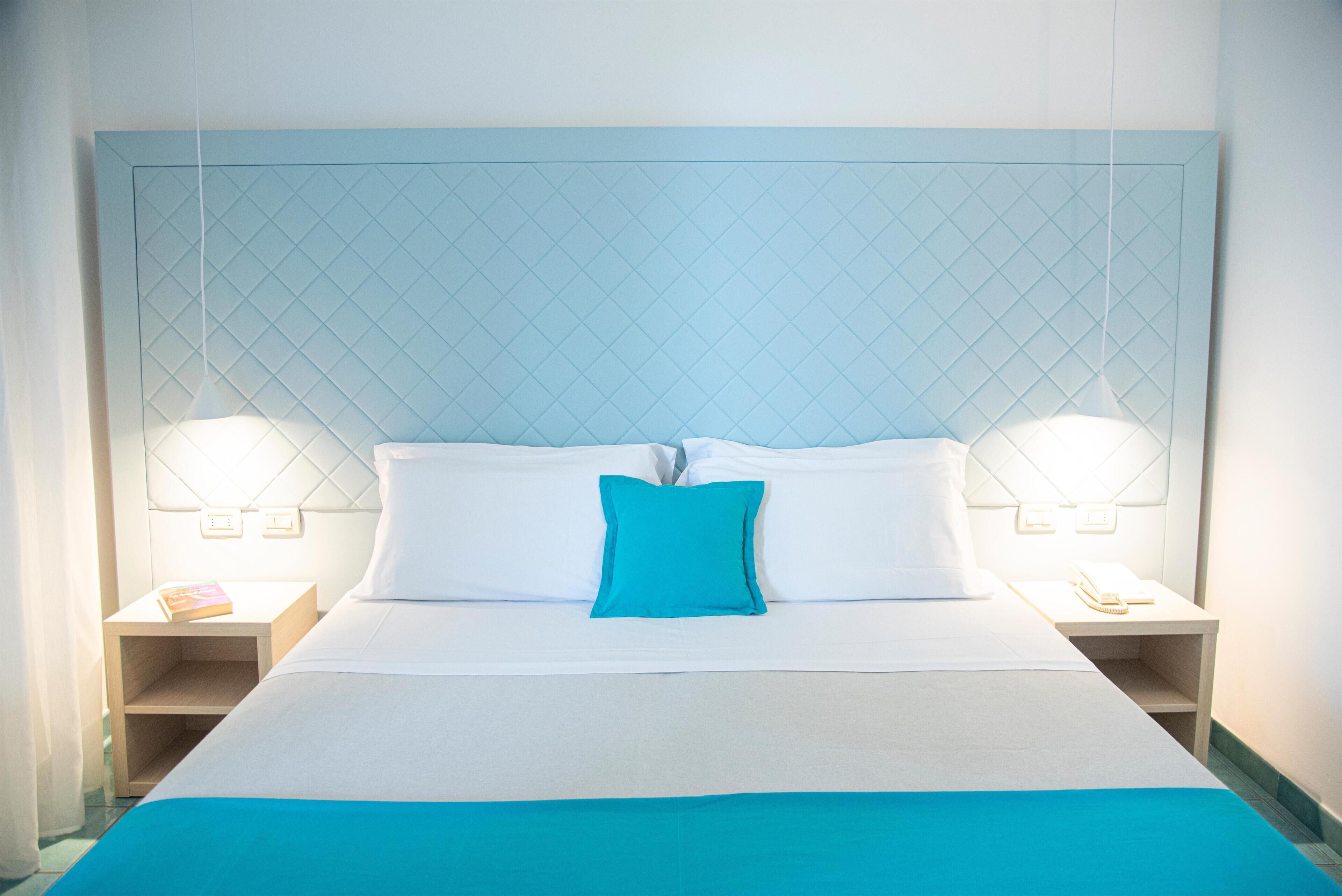 Una cama bien hecha con un cojín azul decorativo, dos lámparas de noche y una mesita sencilla con un teléfono.