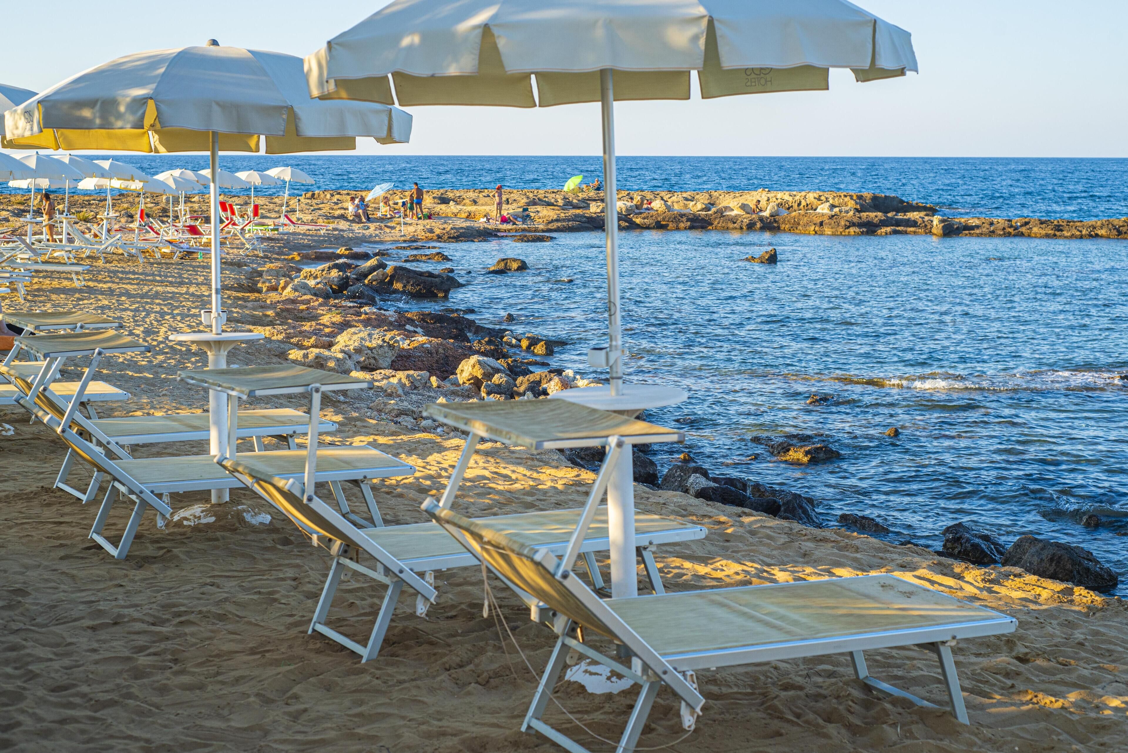 A beach with sun loungers and umbrellas, rocky shoreline, and calm sea.