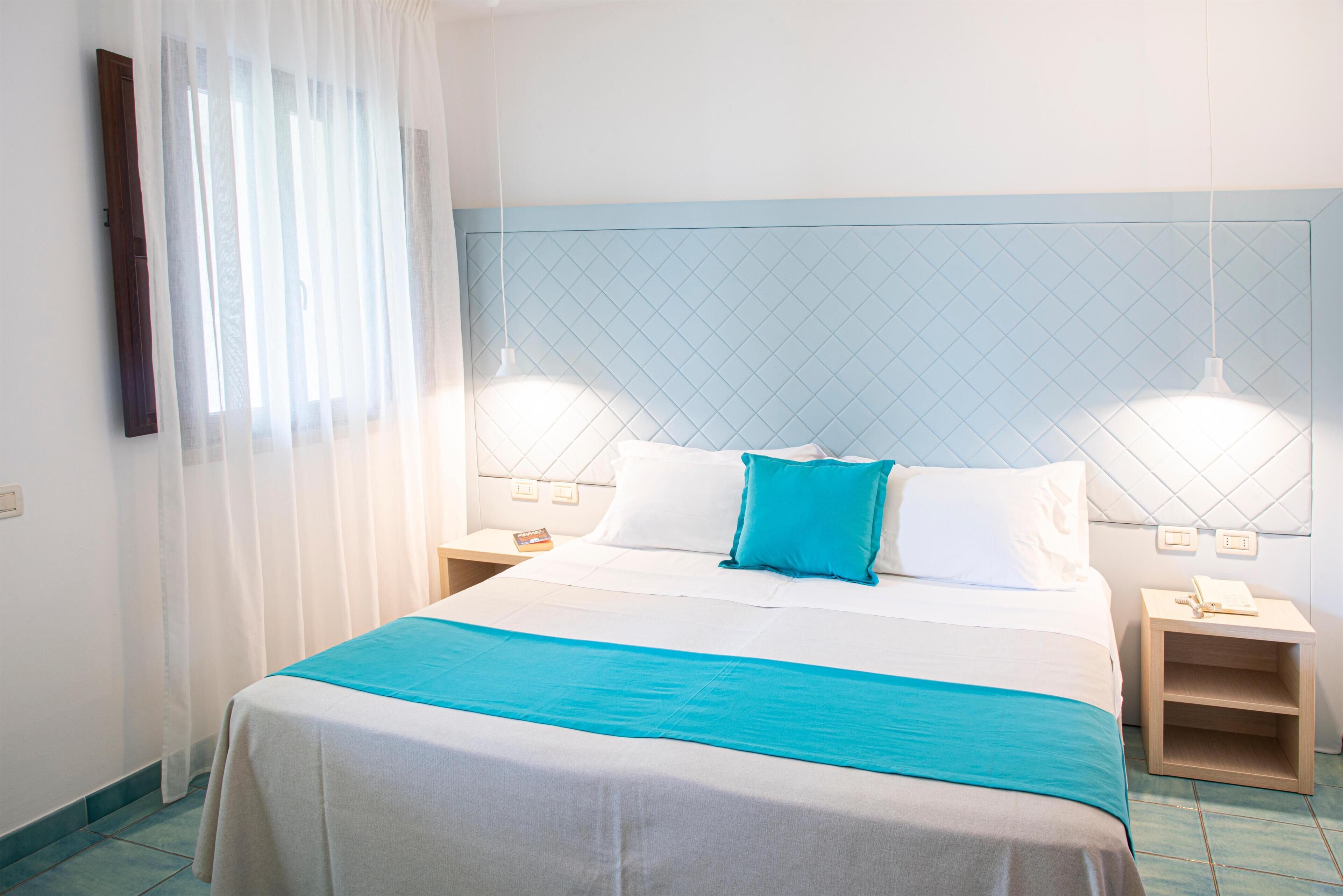 A hotel room with a bed, two bedside tables, a nightstand, a window with sheer curtains, and a wall-mounted light fixture.