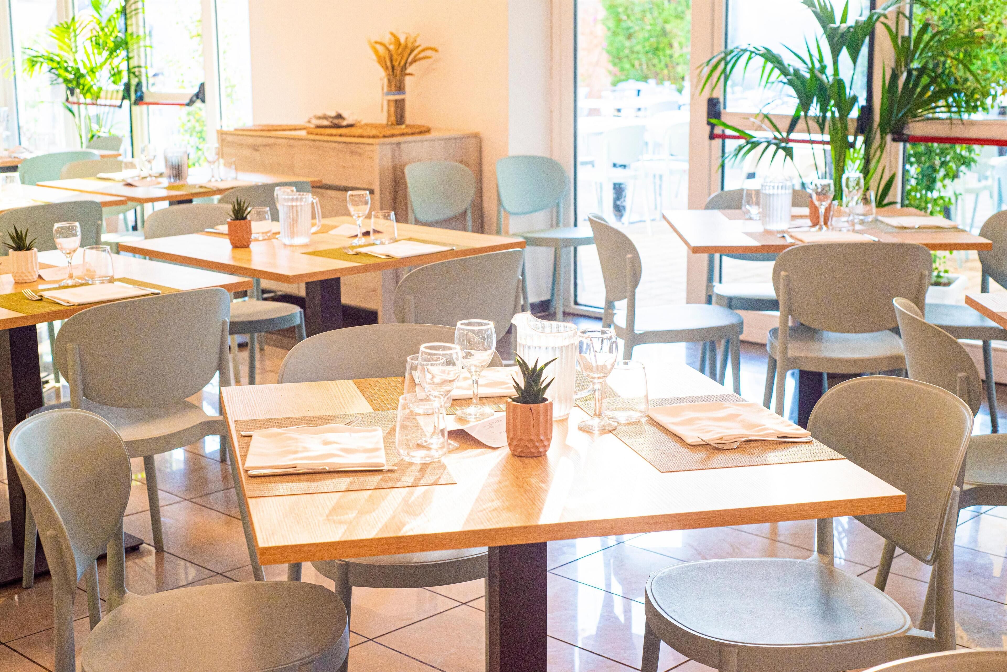 A restaurant with multiple tables and chairs, each set with a placemat, a small plant, and clear glassware.