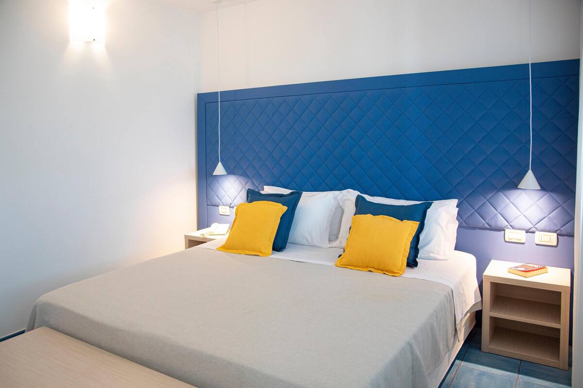 Uma cama bem arrumada com cabeceira azul, duas mesas de cabeceira com abajures e uma prateleira com um livro.