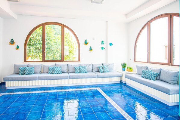 A pool area with a blue tiled floor, a grey sofa with patterned cushions, and large arched windows.