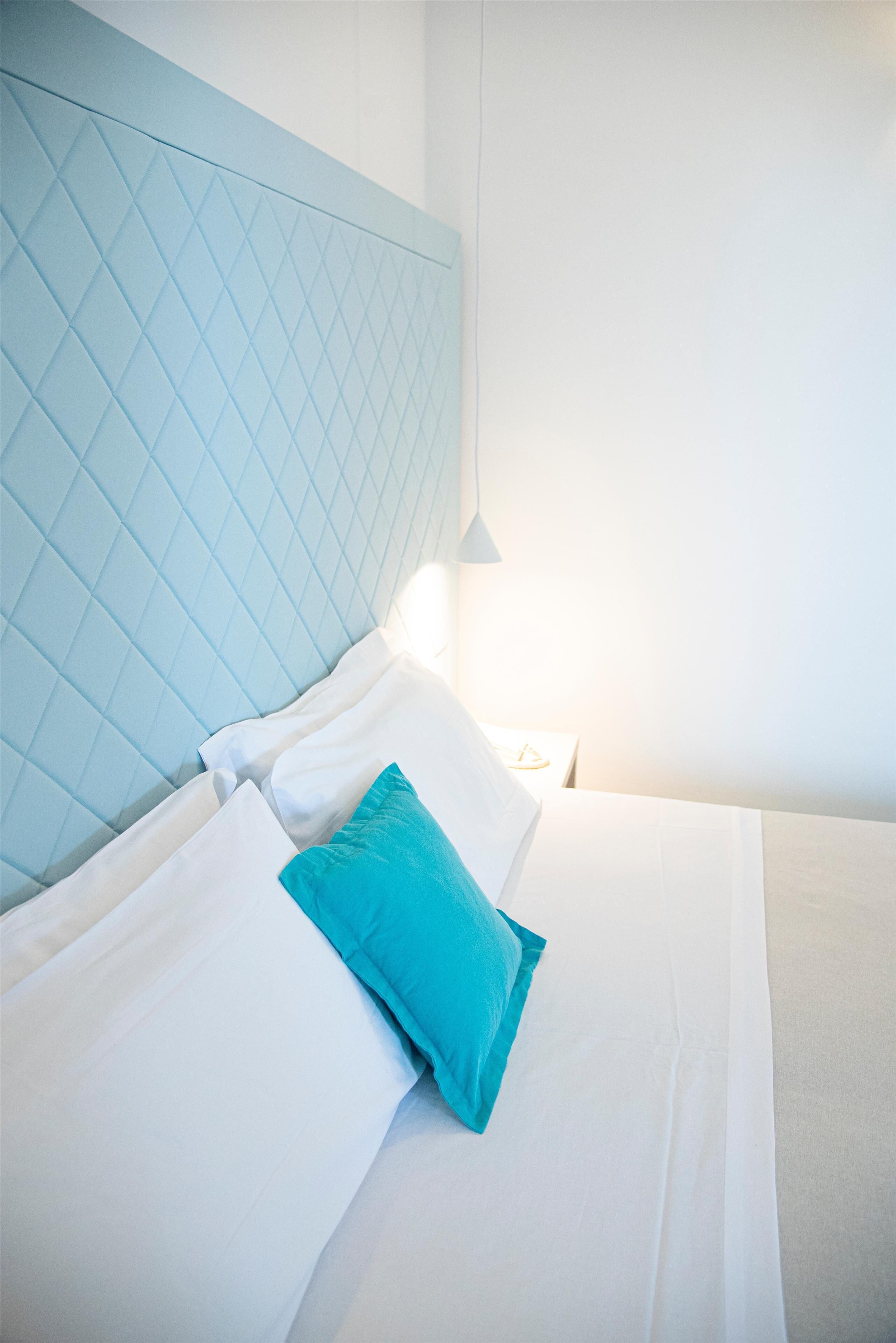 Un lit bien fait, avec des oreillers blancs et turquoise, une tête de lit bleu clair et une lampe de chevet.