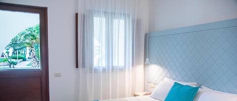 Une chambre d’hôtel avec un lit, une table de chevet, une fenêtre avec des rideaux légers et une vue sur les palmiers à l’extérieur.
