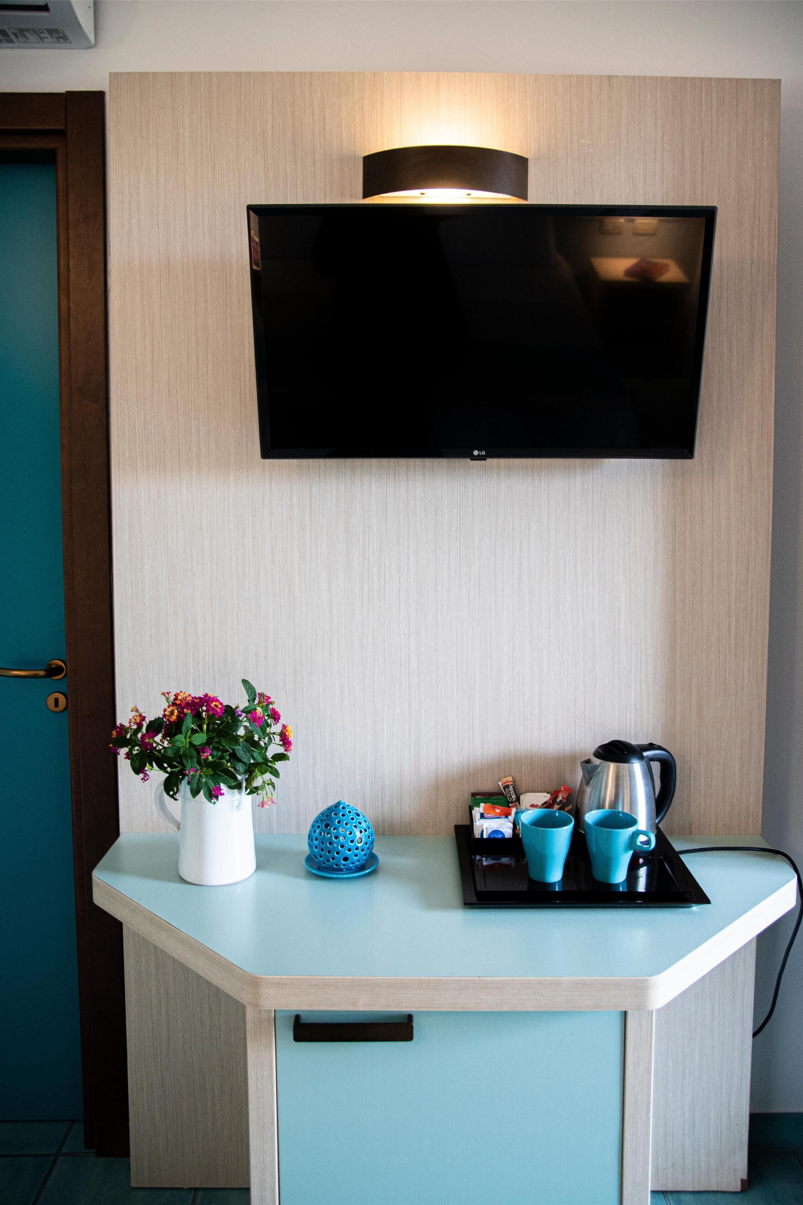 A hotel room with a flat-screen TV mounted on the wall, a small table with a vase of flowers, a blue decorative item, and a tray with a coffee pot and cups.
