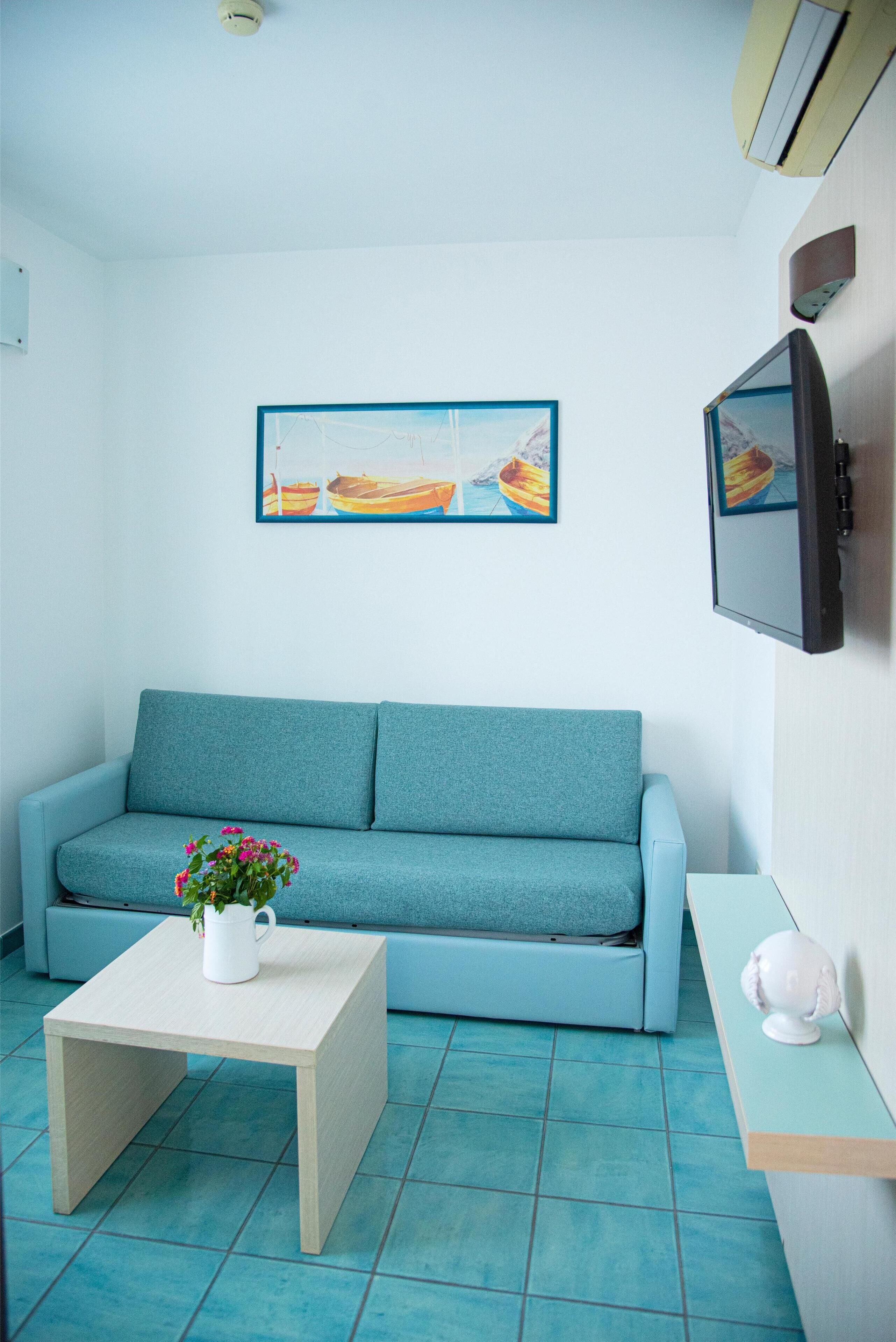 A room with a teal sofa, a small wooden table with a flower vase, a flat-screen TV mounted on the wall, and a painting of boats on the wall.