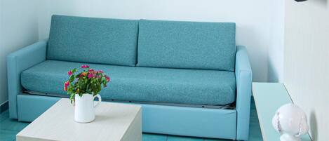 A room with a teal sofa, a small wooden table with a flower vase, a flat-screen TV mounted on the wall, and a painting of boats on the wall.