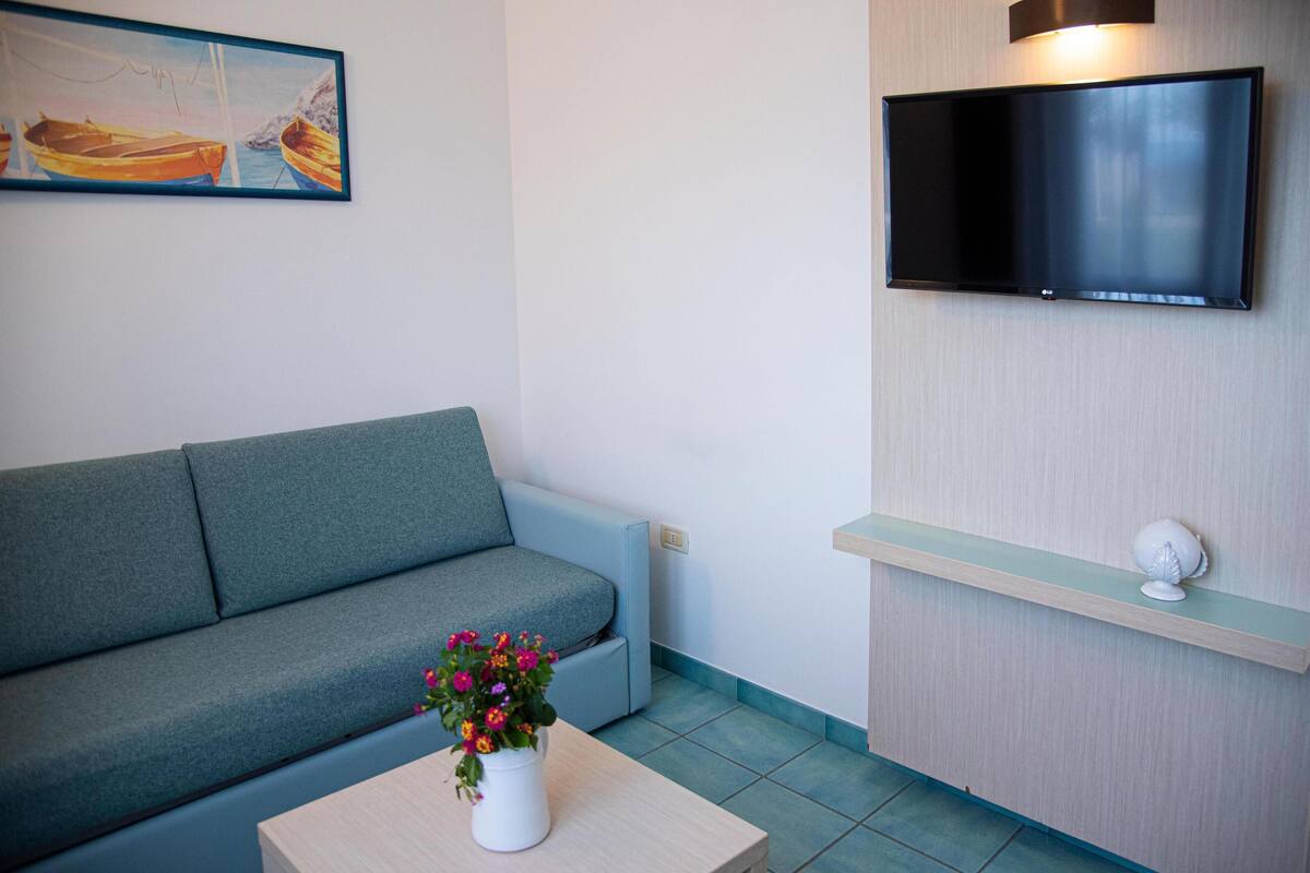Uma sala de estar moderna com um sofá cinzento, uma pequena mesa de centro de madeira com um vaso de flores, uma TV de tela plana montada na parede e uma estante clara com um objeto decorativo.