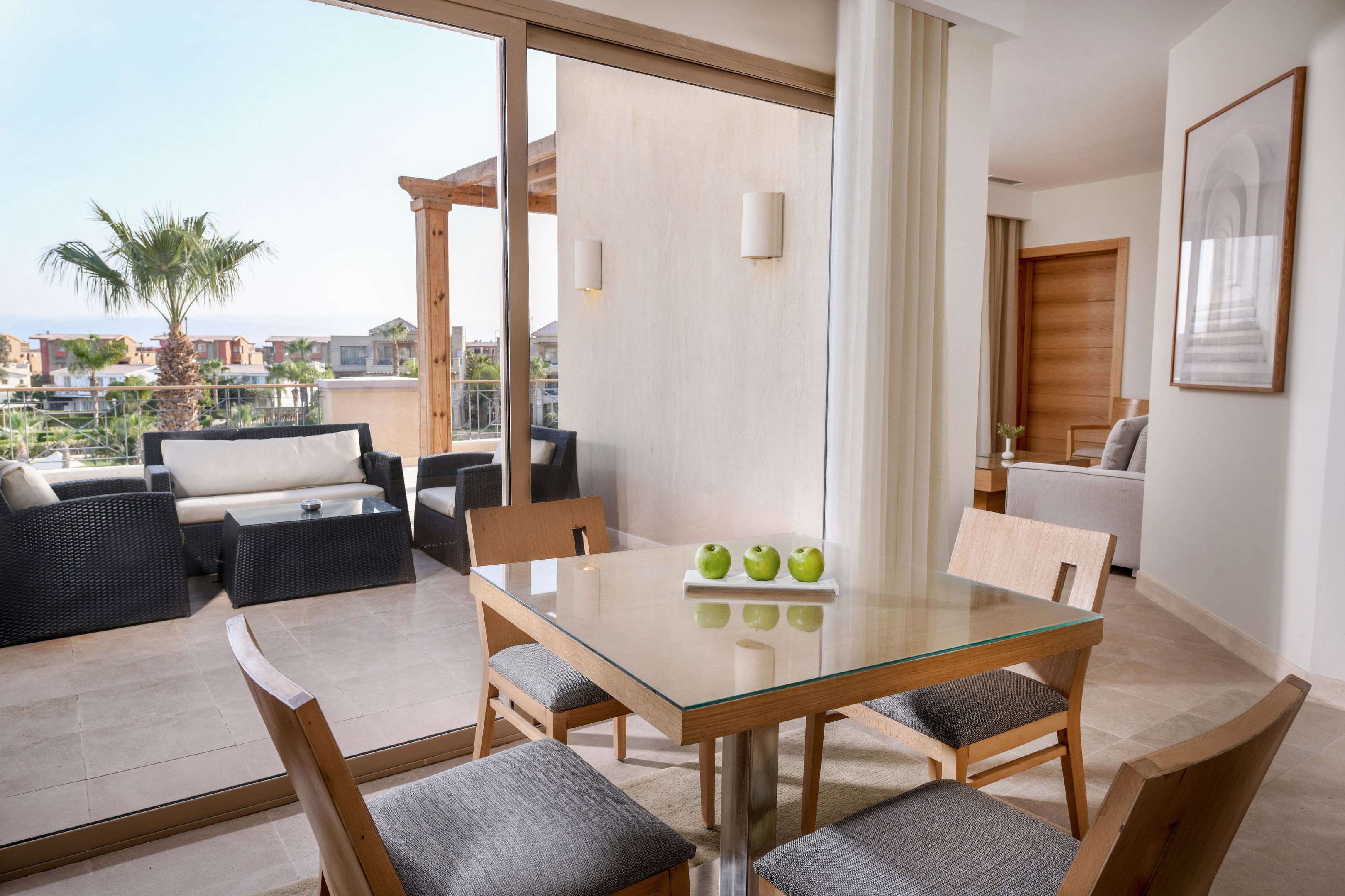 A modern dining area with a glass table, four chairs, and a view of palm trees and buildings.