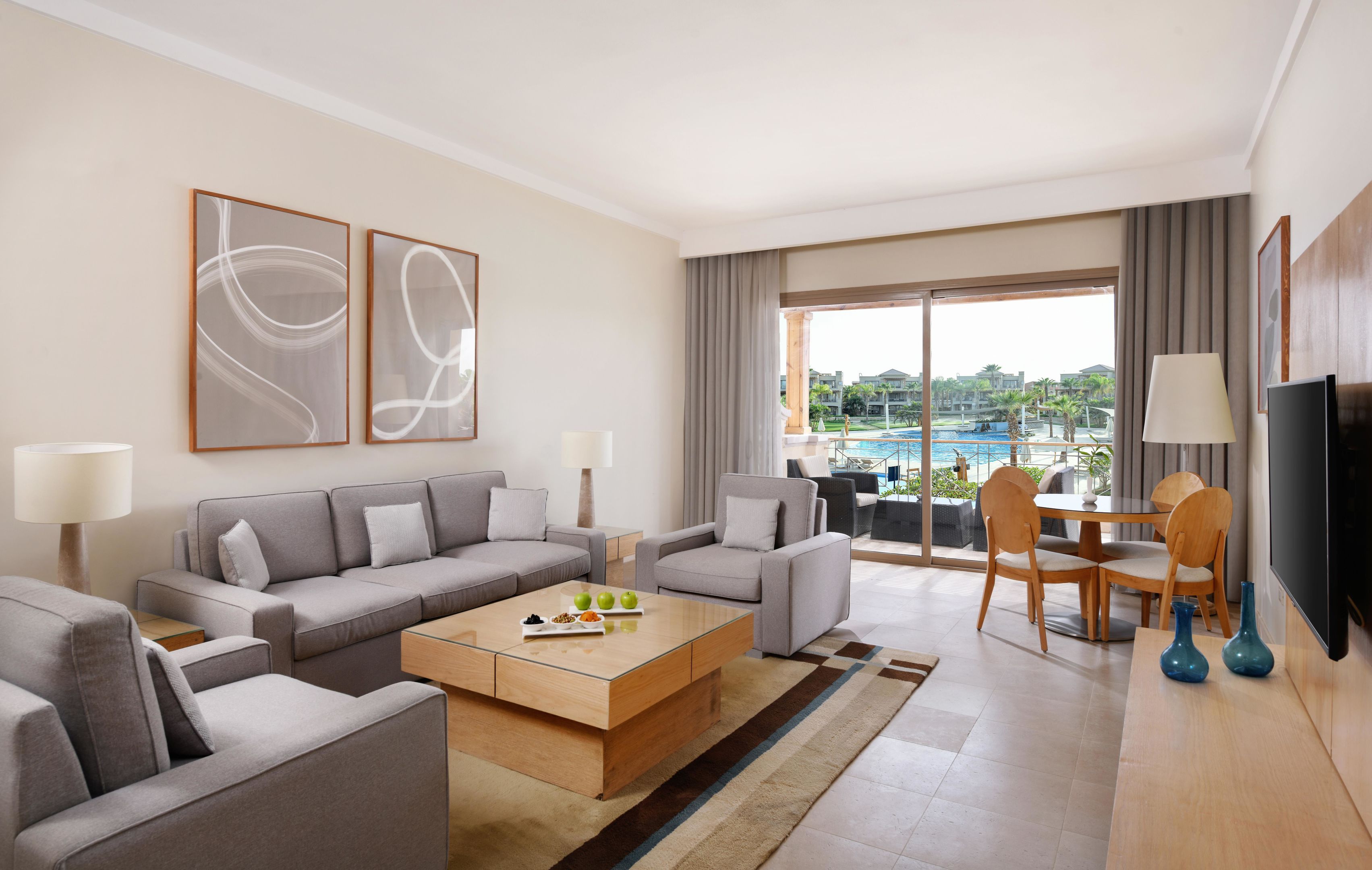 Una habitación de hotel moderna con sofá, mesa de centro, mesa de comedor con sillas, televisión y un gran ventanal con vistas a una piscina y edificios.