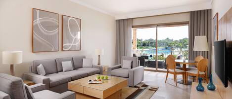 A modern hotel room with a sofa set, a coffee table, a dining table with chairs, a TV, and a large window with a view of a pool and buildings.