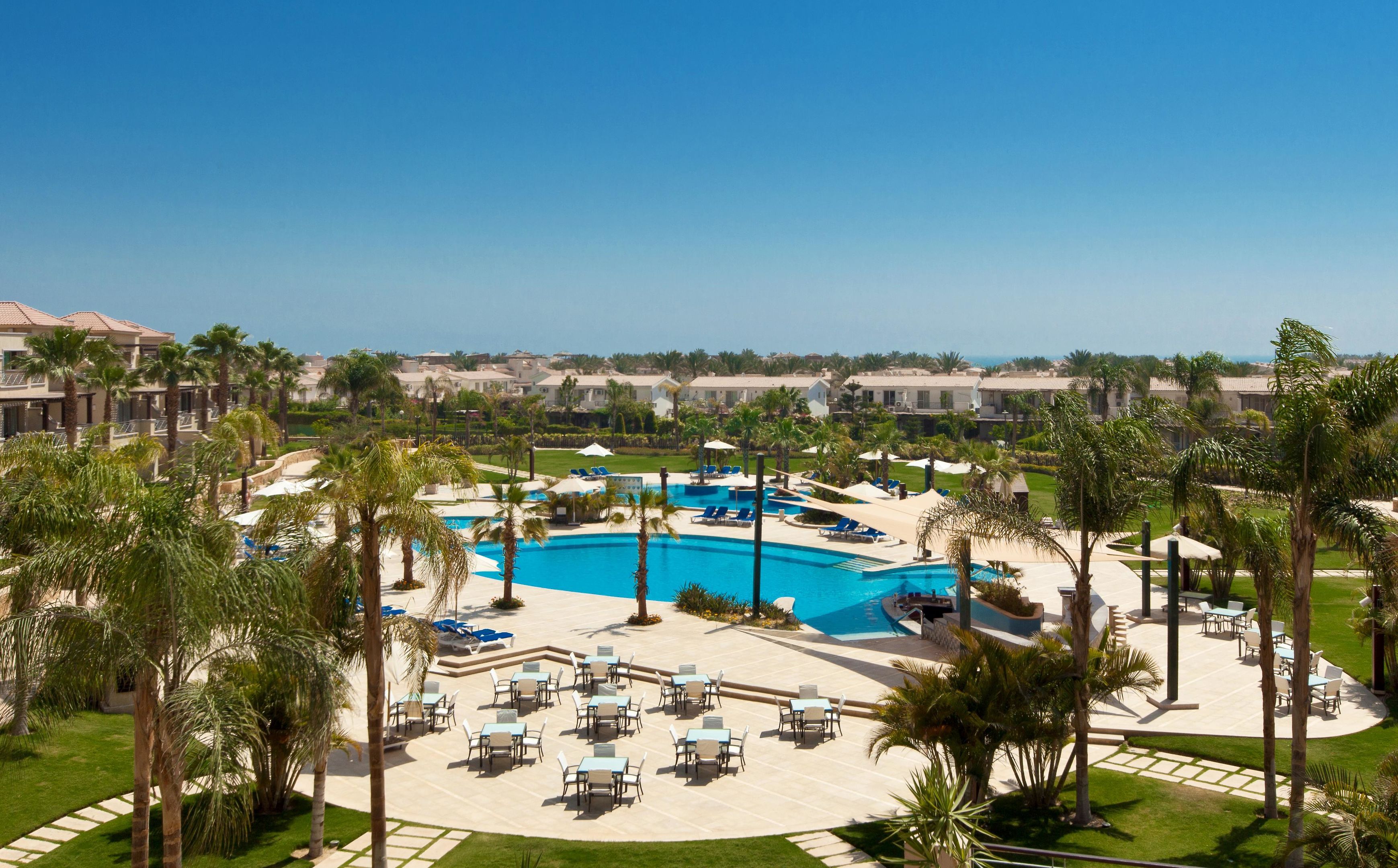 A resort with a central swimming pool, outdoor seating, and palm trees.
