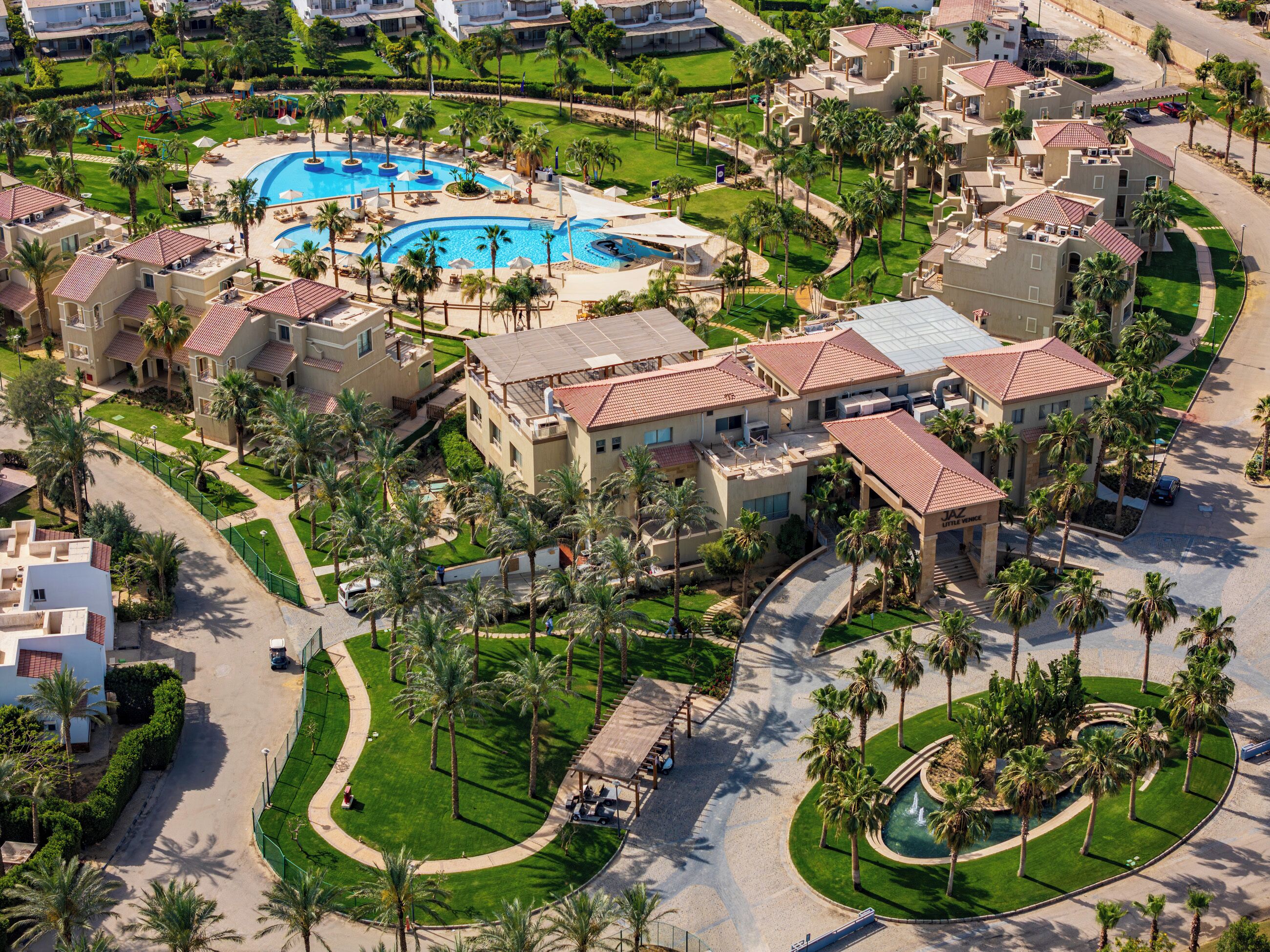 Luchtbeeld van een resort met meerdere gebouwen, een centraal zwembad en omliggende palmbomen.