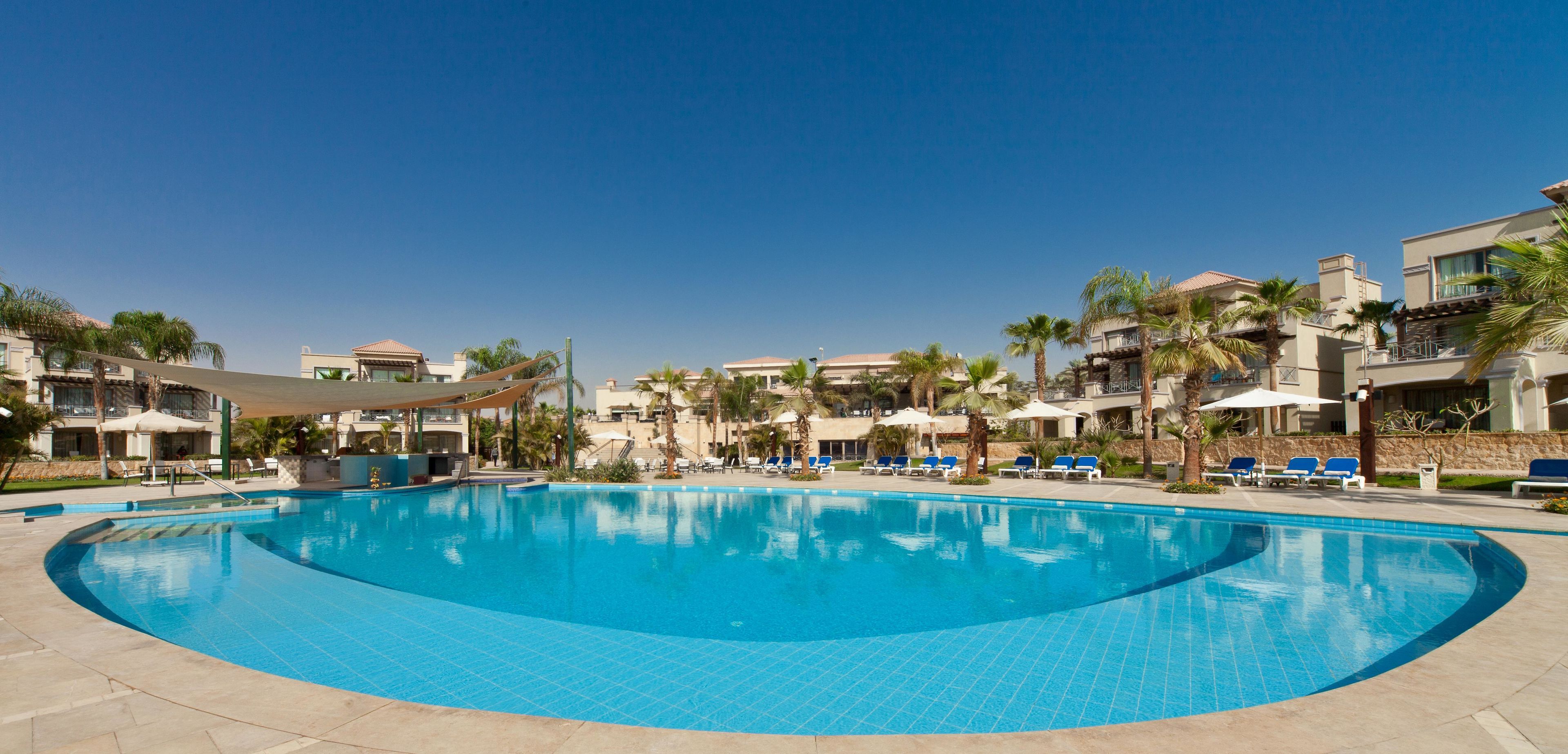 A large swimming pool with lounge chairs and umbrellas, surrounded by buildings with balconies and palm trees.