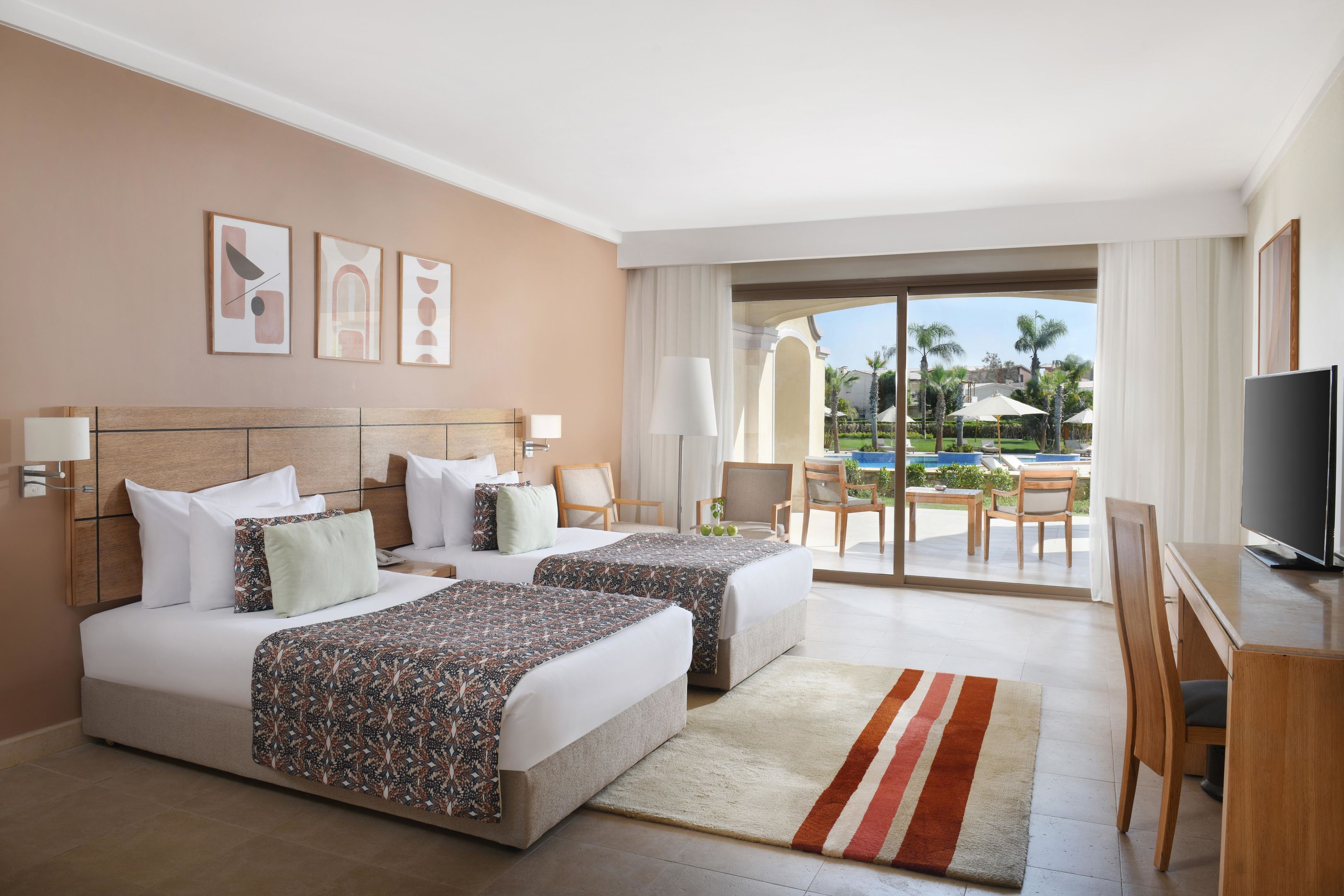 Une chambre d’hôtel avec deux lits, une télévision, un bureau et une vue sur la piscine et les palmiers.