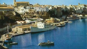Un pueblo costero con un barco blanco grande amarrado en el puerto, embarcaciones más pequeñas y edificios a lo largo del paseo marítimo.