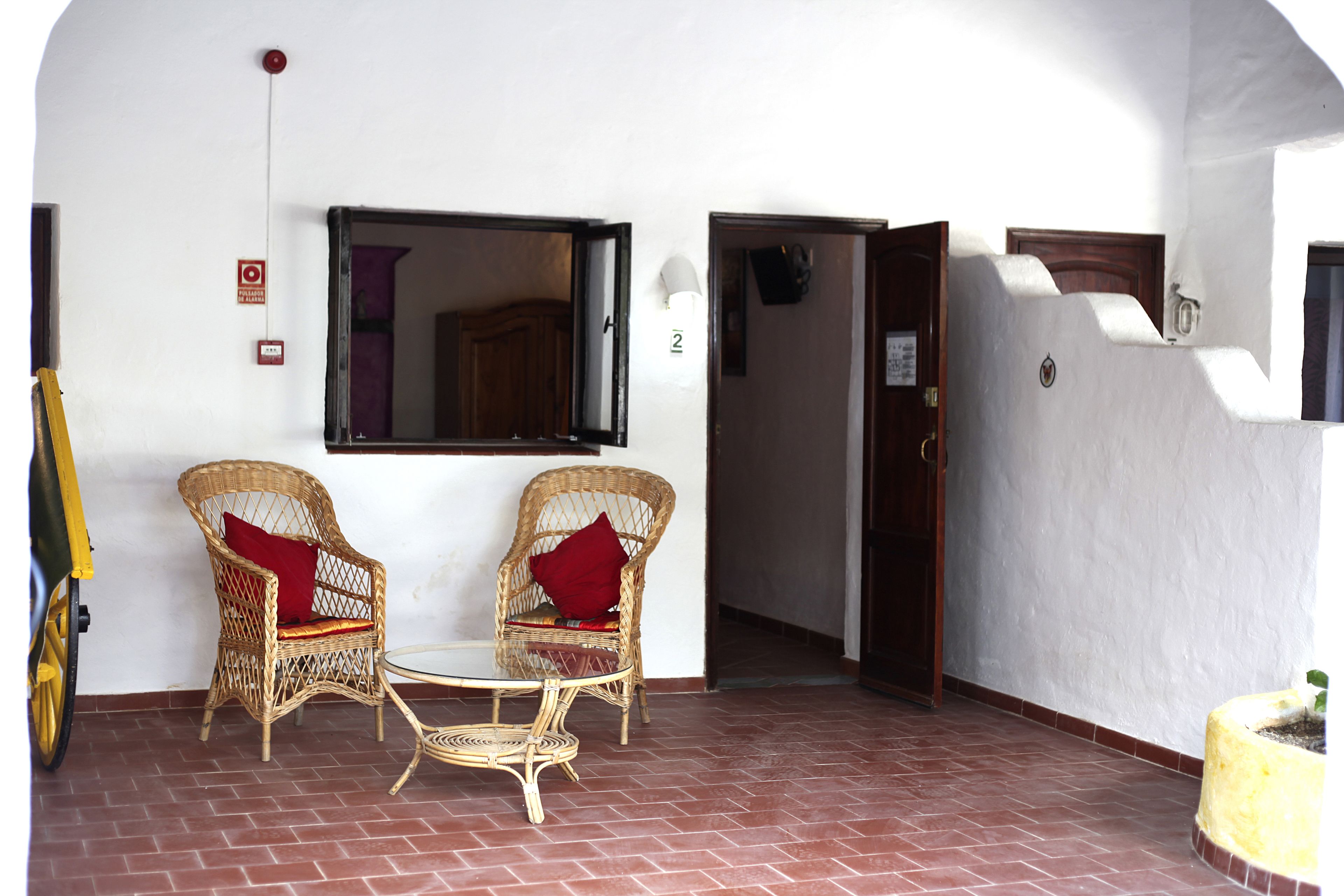 Une pièce avec deux fauteuils en osier, une petite table ronde et un miroir au mur. Il y a une porte qui mène à une autre pièce et un vélo jaune à l’extérieur.