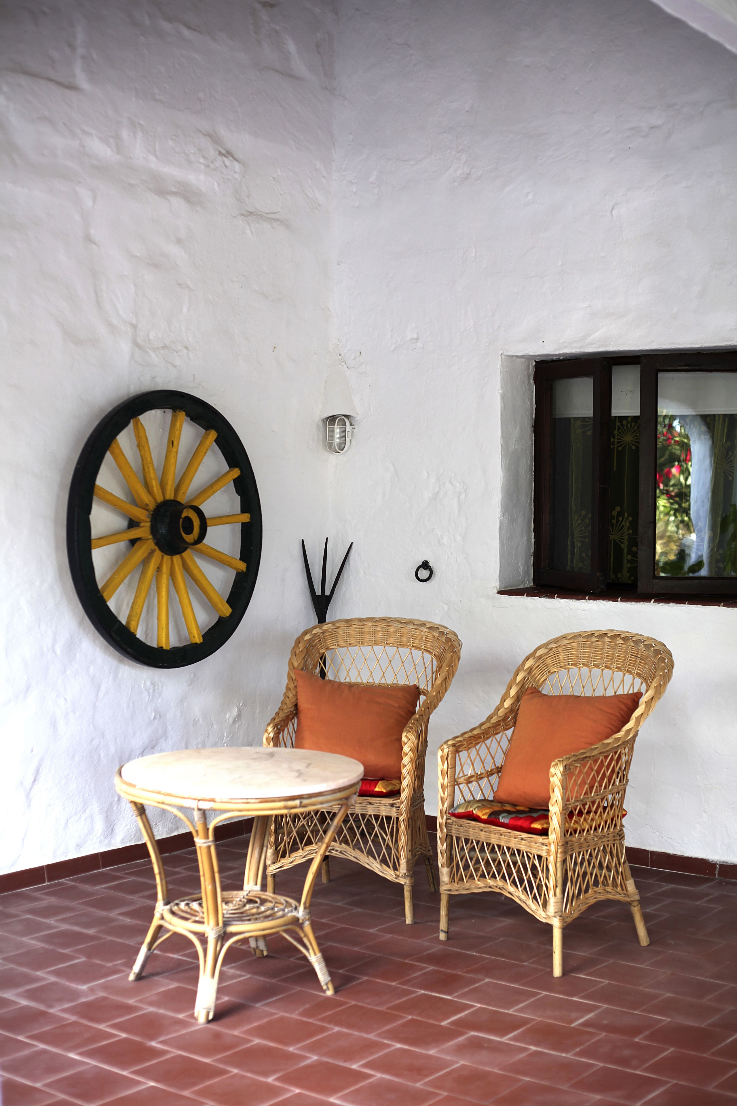 Una habitación con dos sillones de mimbre y una pequeña mesa redonda, una gran rueda decorativa en la pared y una ventana con vistas a la vegetación.