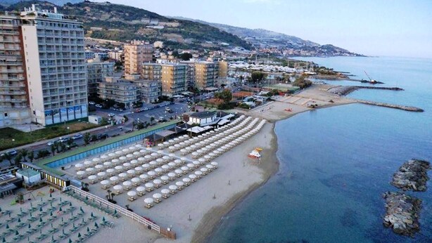 Un hotel frente al mar con varias edificaciones, una playa con sombrillas y vistas al océano y colinas al fondo.
