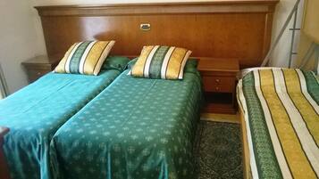 A room with two beds, each with green bedding and striped pillows. There is a wooden headboard and a small bedside table with a lamp.
