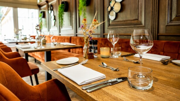 A restaurant with wooden tables, orange upholstered booths, and a candle centerpiece. - HOTEL HERIKERBERG (Markelo)