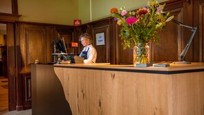 A reception desk with a wooden counter, a computer, and a vase with flowers. - HOTEL HERIKERBERG (Markelo)