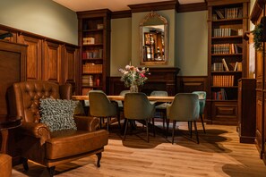 A library with a leather armchair, a wooden table, and bookshelves. - HOTEL HERIKERBERG (Markelo)