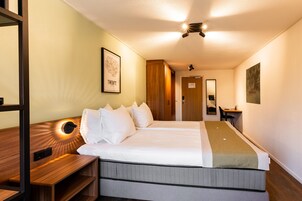 Quarto de hotel com uma cama grande, um banquinho de madeira, uma escrivaninha e um espelho.