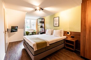A neatly arranged bedroom with a large bed, a bedside table, a bookshelf, and a window providing natural light.