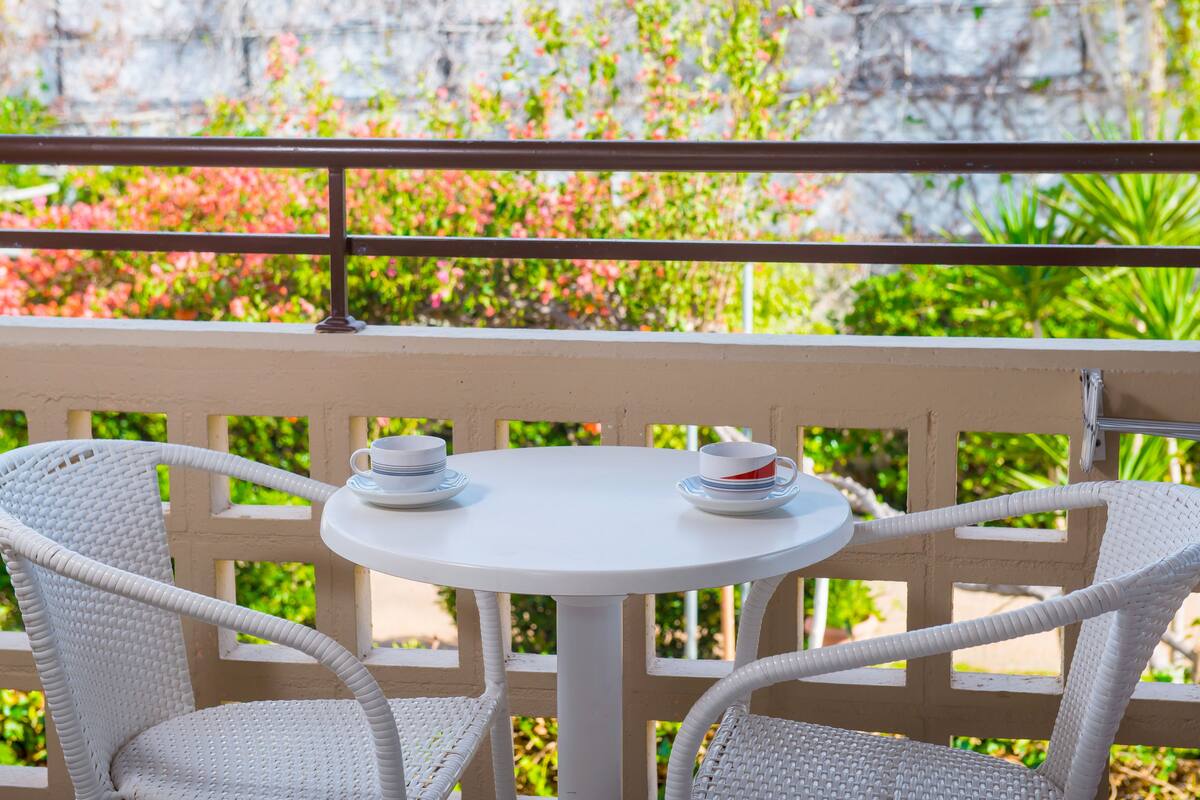 A white wicker table with two cups and saucers, set against a balcony railing with greenery in the background.