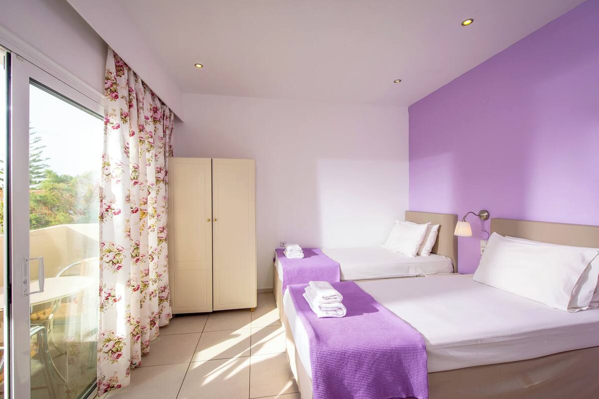 A hotel room with two beds, a wardrobe, a bathroom, and a balcony with curtains.