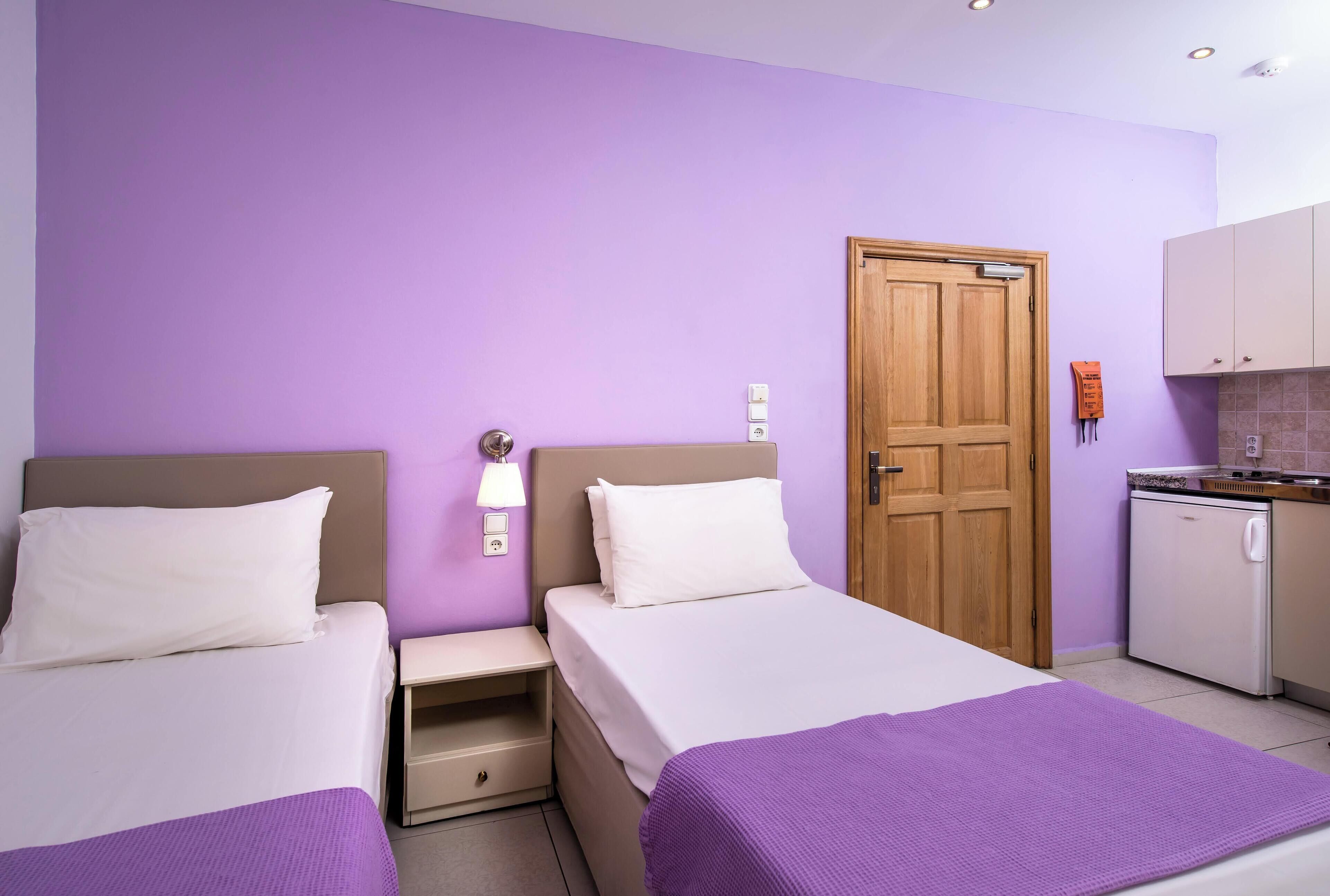 A hotel room with two beds, a wooden door, a small kitchenette, and a purple wall.