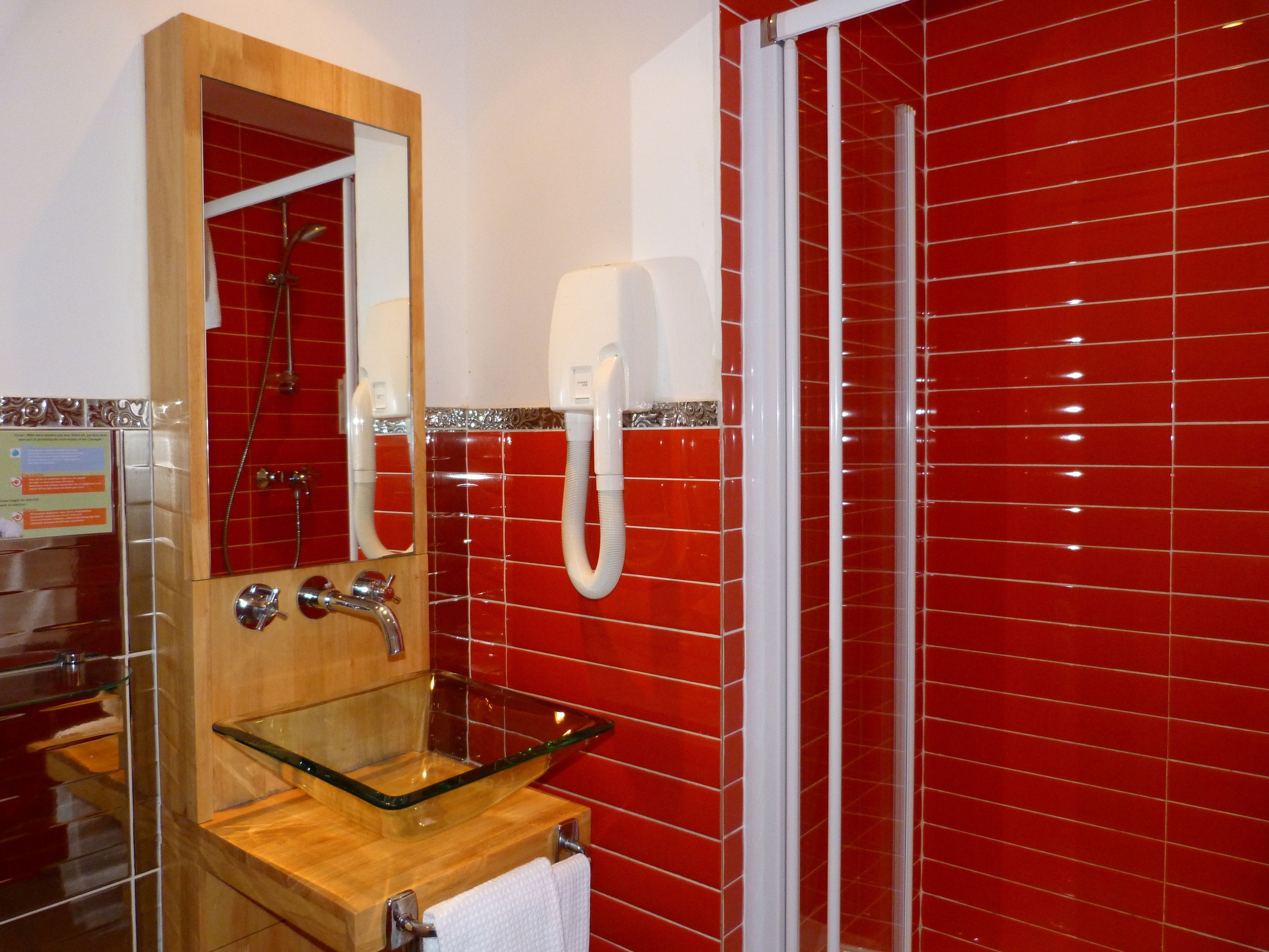 Un bagno con un lavandino in vetro, uno specchio, un soffione doccia a mano e una parete rivestita di piastrelle rosse.