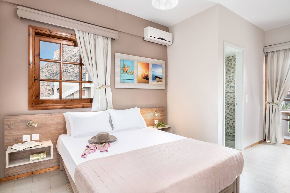 Une chambre avec un grand lit, une tête de lit en bois, une table de chevet, une fenêtre avec des rideaux et un climatiseur fixé au mur.