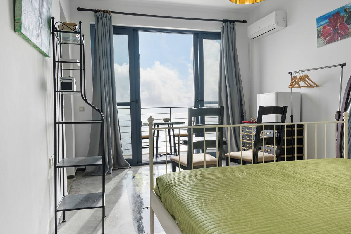 A bedroom with a bed, a balcony with chairs, a metal shelf unit, and a painting on the wall.