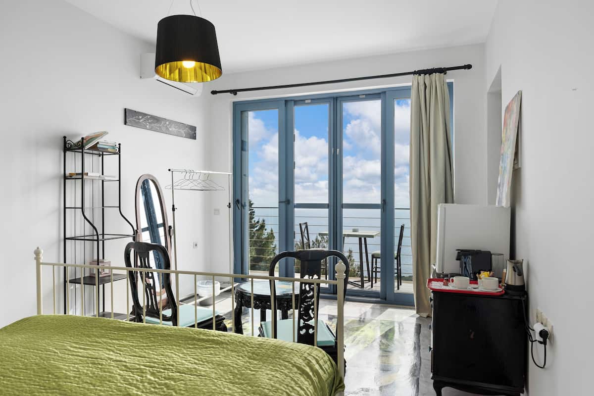 A bedroom with a bed, a desk, a chair, a table, a hanging light fixture, and a view of the sky through a large window.