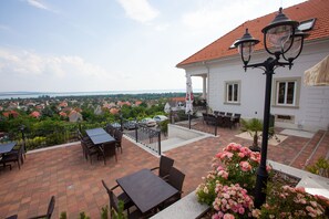 A terrace with outdoor seating, a view of the city, and a building with a red roof. - Zenit Hotel Balaton (Vonyarcvashegy)