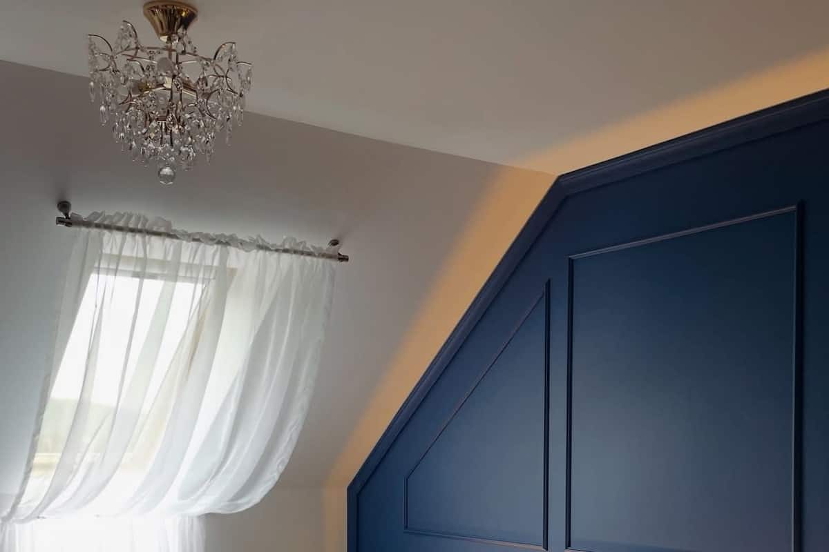 A hotel room with a bed, a chandelier, a window with curtains, and a wall-mounted light.