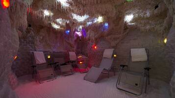 A cave with a seating area featuring lounge chairs and a small table, illuminated by colorful lights.