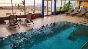 An indoor swimming pool with lounge chairs and large windows offering a view of the beach. - Zenit Hotel Balaton (Vonyarcvashegy)