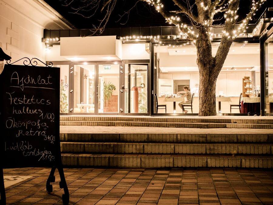 A nighttime view of a restaurant with outdoor seating, a signboard with menu items, and a tree decorated with lights.