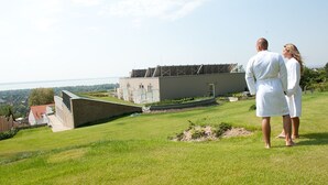 A couple standing on a grassy hill overlooking a modern building with a sloped roof and a view of the sea. - Zenit Hotel Balaton (Vonyarcvashegy)